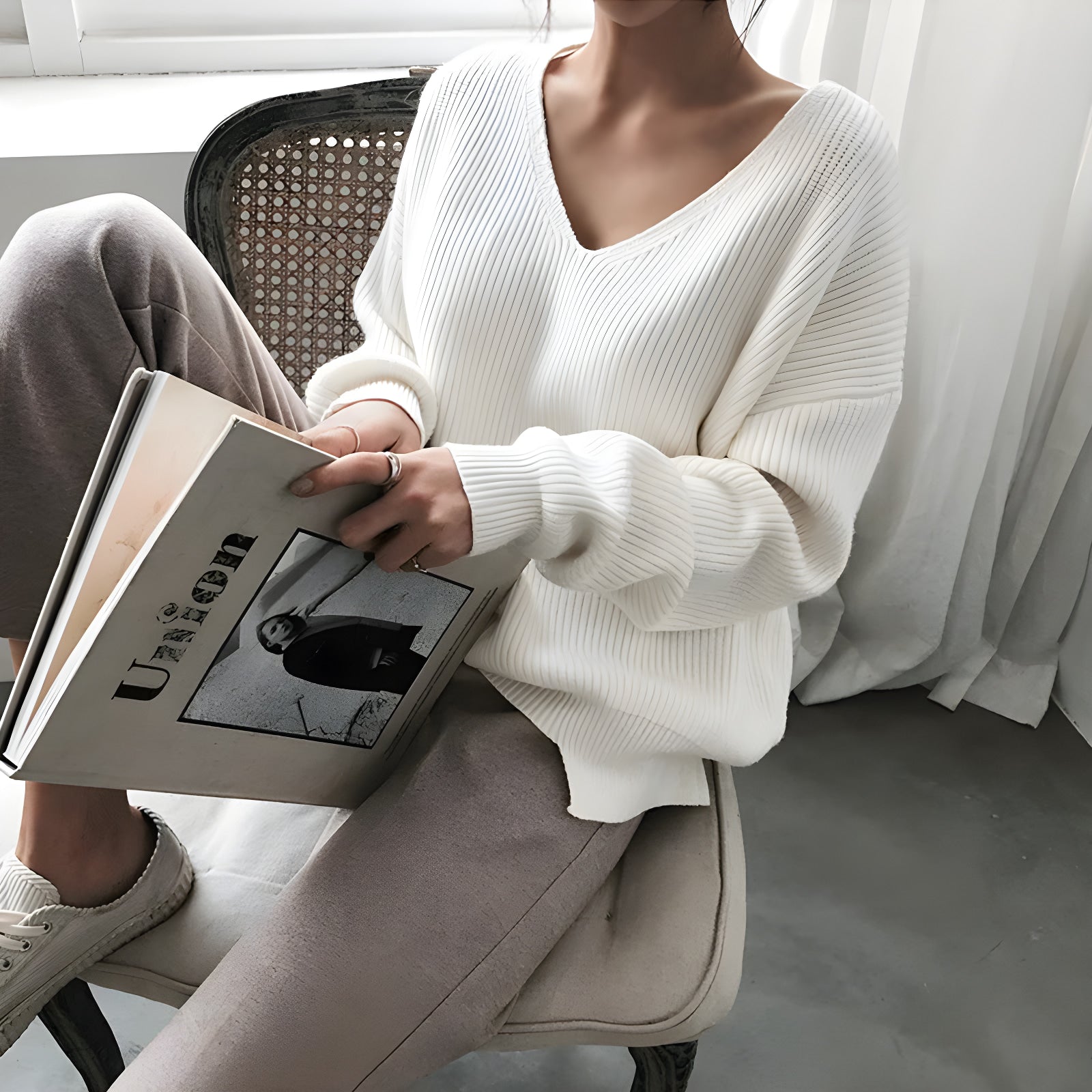 Person wearing a white sweater and beige pants holding a book indoors.  #Color_White